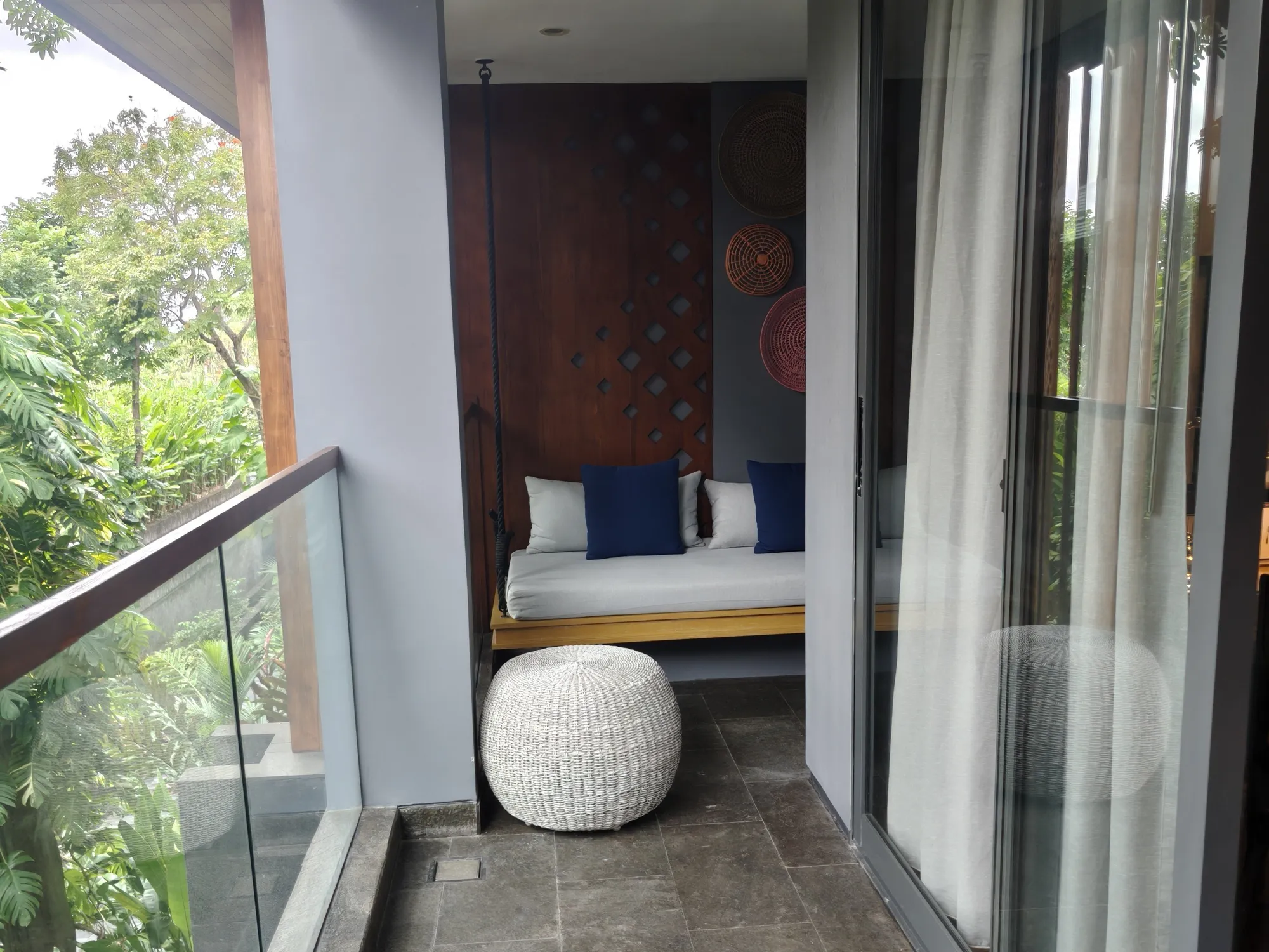 A balcony with a hanging daybed, a woven pouf and a glass rail looking out over the gardens