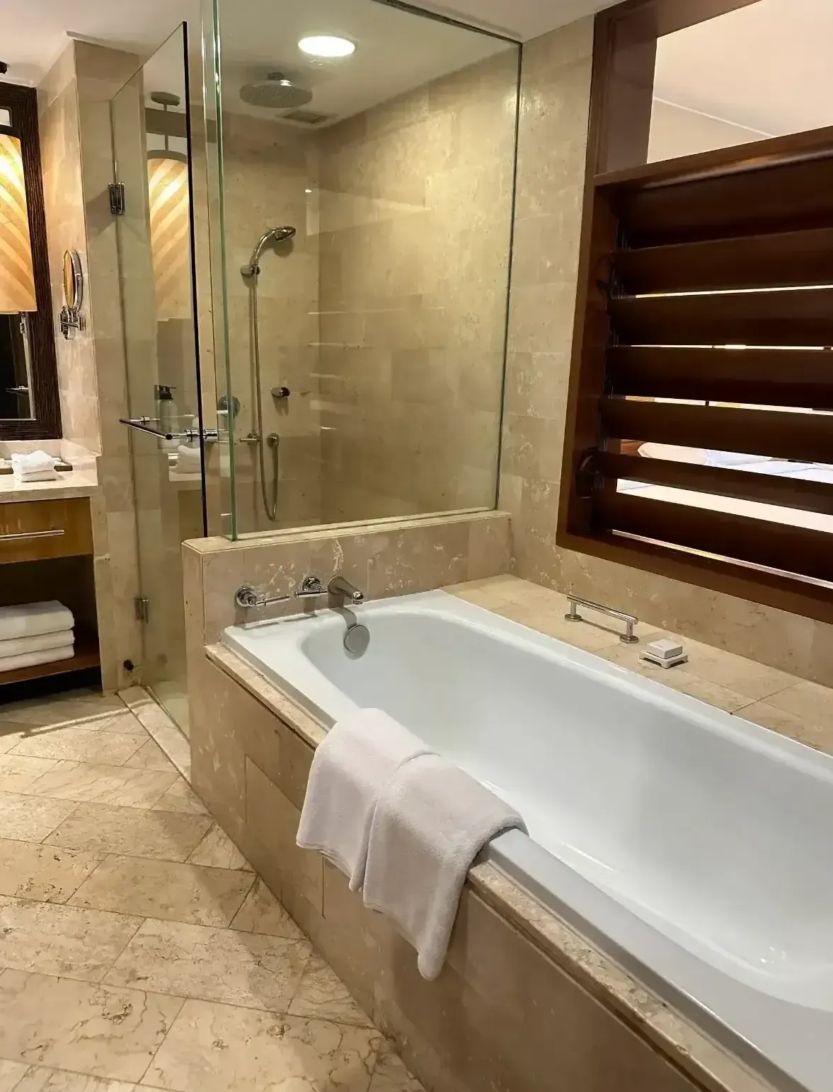A Grand Hyatt bathroom with a marble-clad bathtub, a glass rain shower and wooden shutters opening to the bedroom