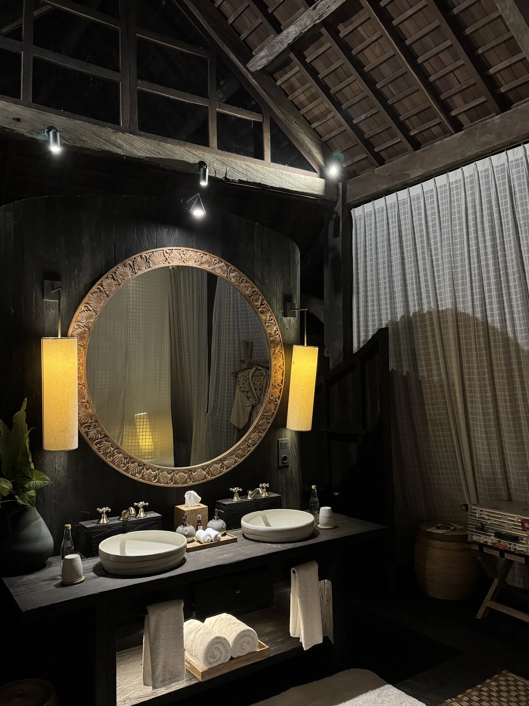Twin basins under a round carved mirror and paper lamps, in a dark timber bathroom with a peaked roof