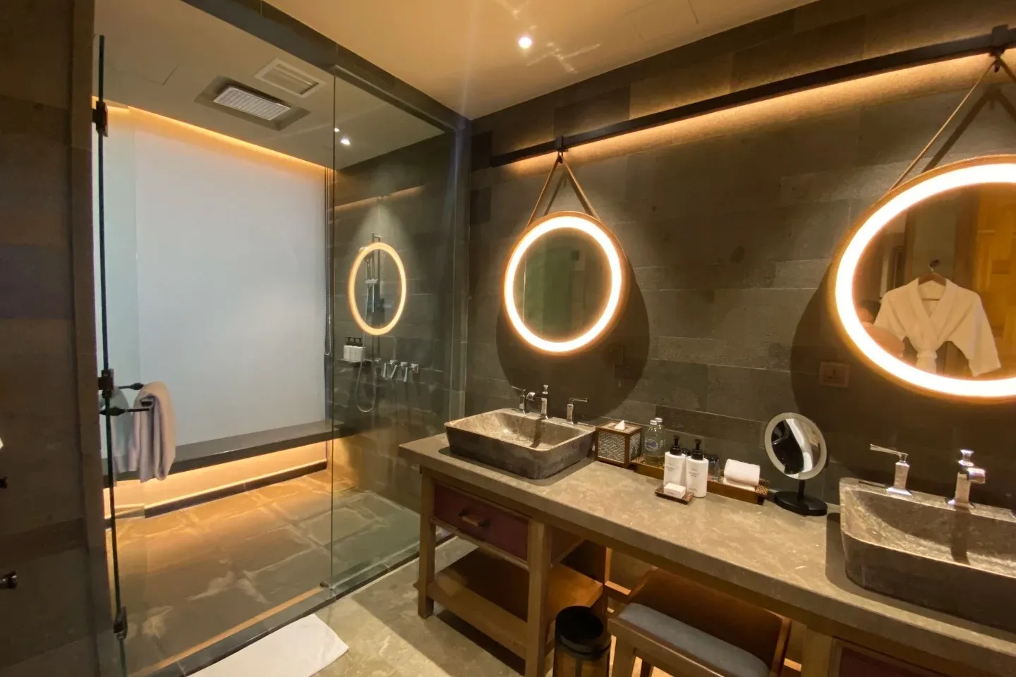An Andaz bathroom with twin stone basins under round backlit mirrors and a glass rain shower