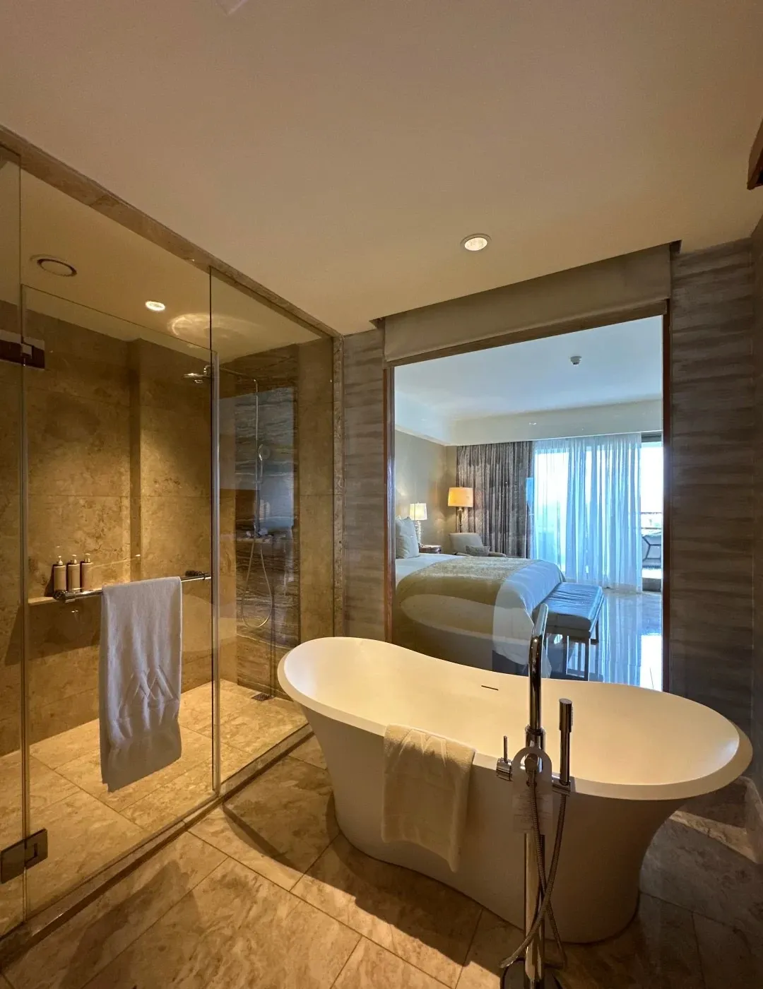 A freestanding bathtub beside a glass shower, with the bedroom seen through an interior window
