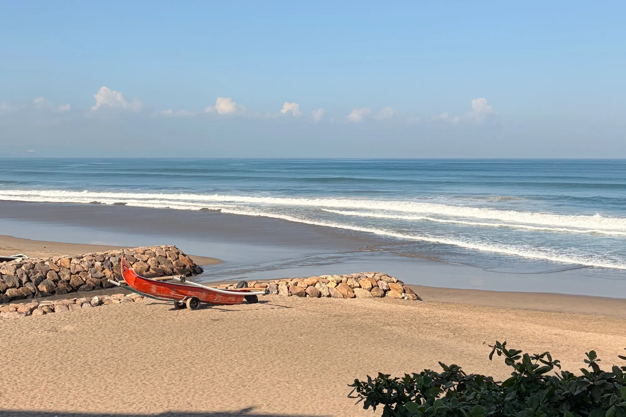 A red fishing boat on the sand by a rock groyne, with surf rolling in on the beach in front of Alila Seminyak