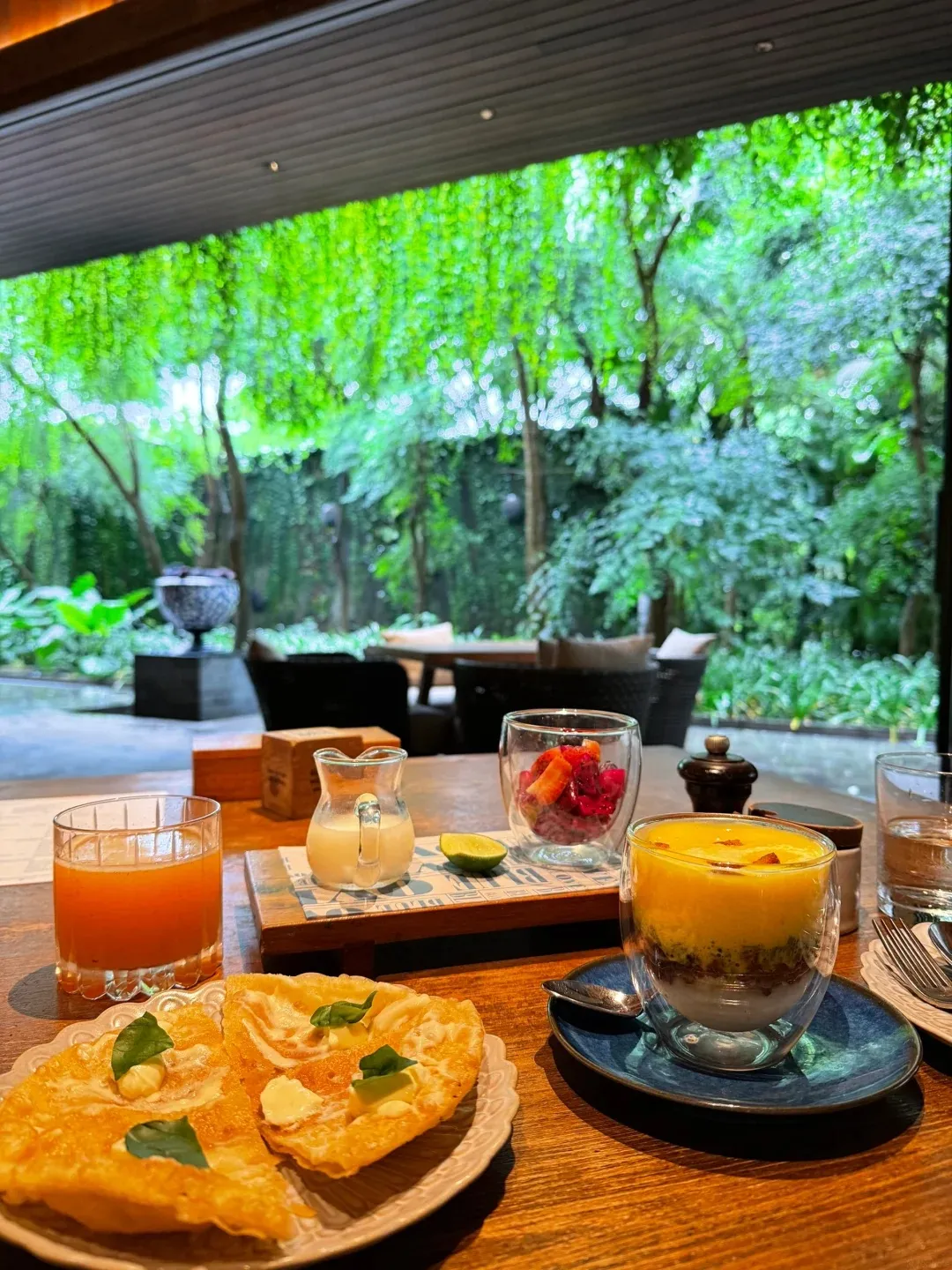 Breakfast of fresh juice, a parfait and flatbread on a table looking out to the garden