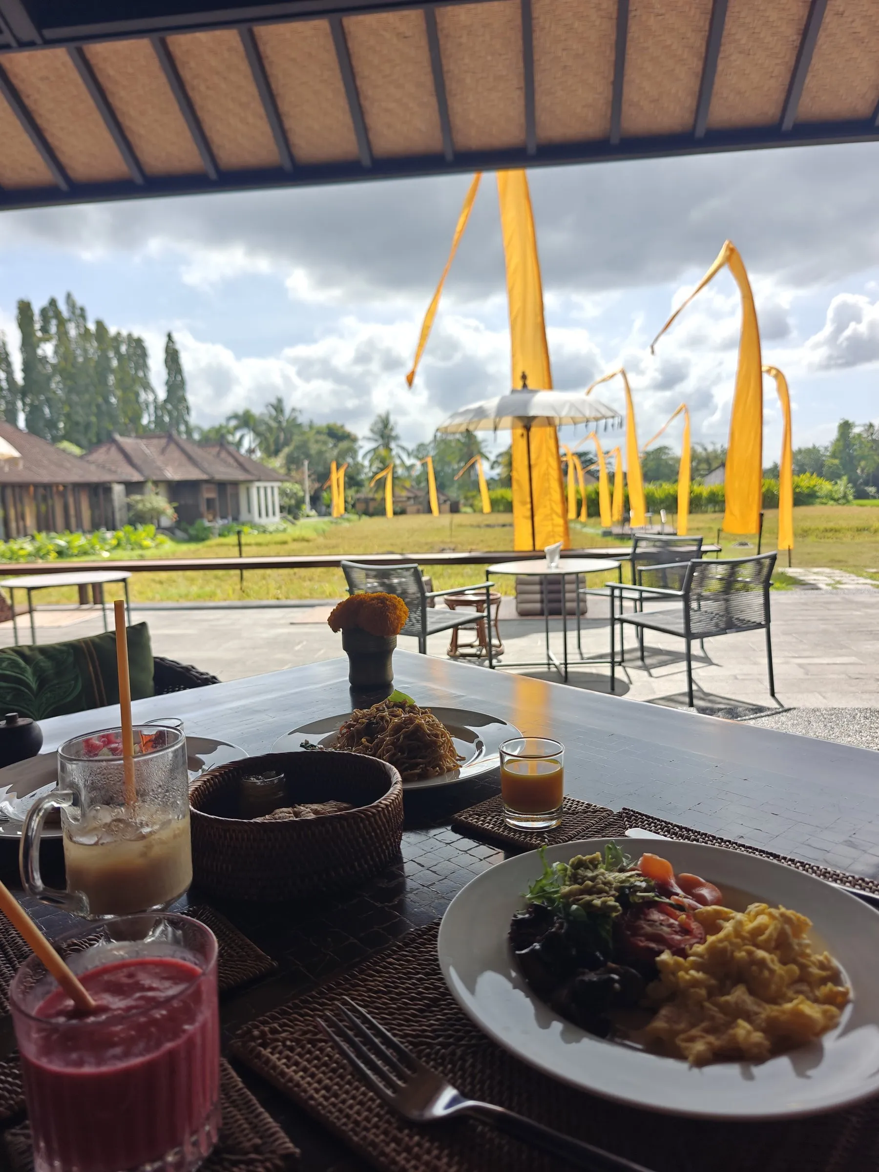 Breakfast eggs, juices and a smoothie on a covered terrace, looking out to the paddy and tall bamboo penjor poles
