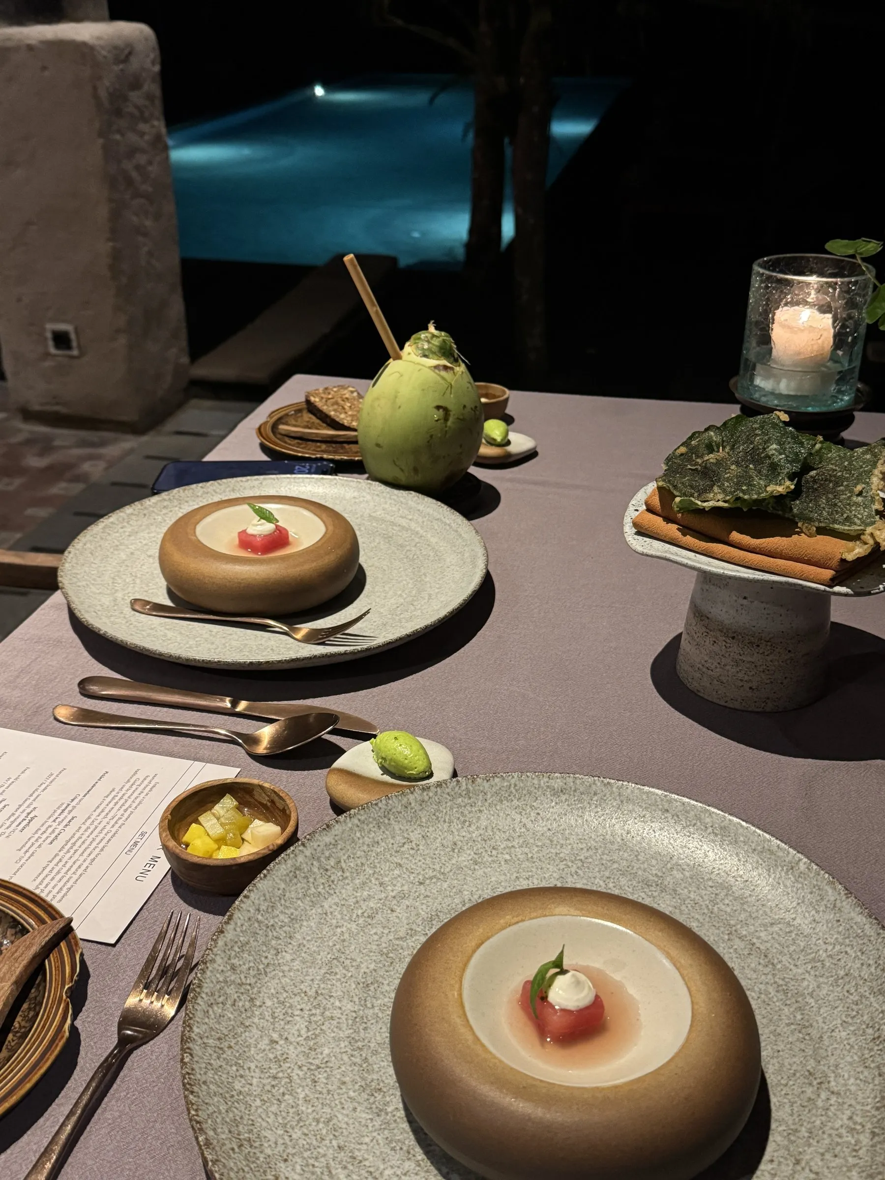 A dinner table by candlelight with a fresh coconut and small plated courses, beside the pool lit up at night