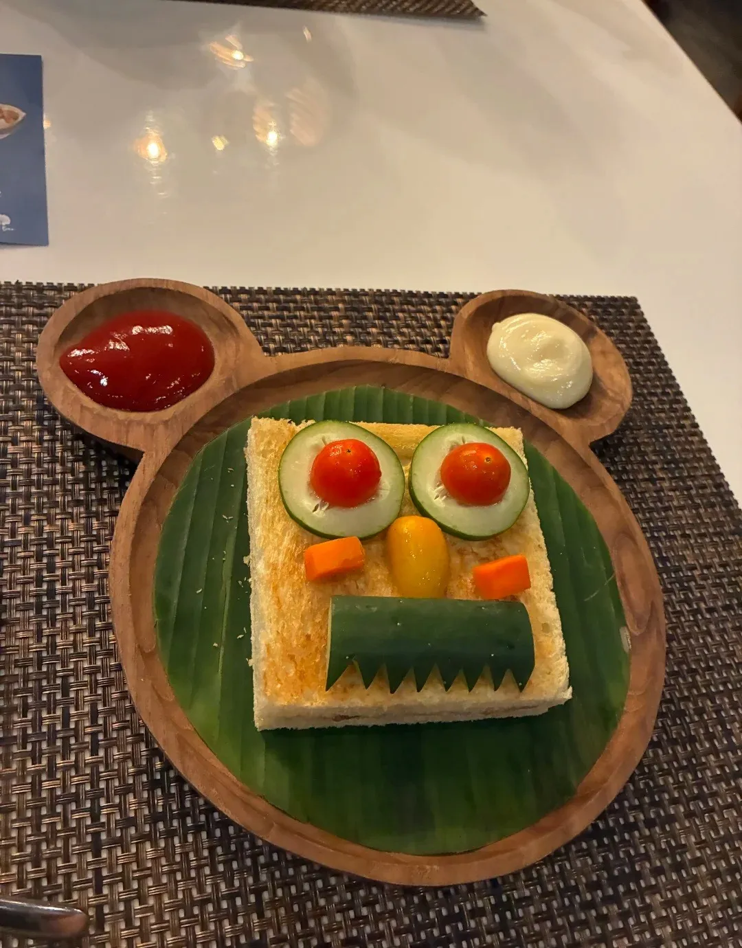 A kids breakfast of toast made into a face, with tomato eyes and a cucumber mouth