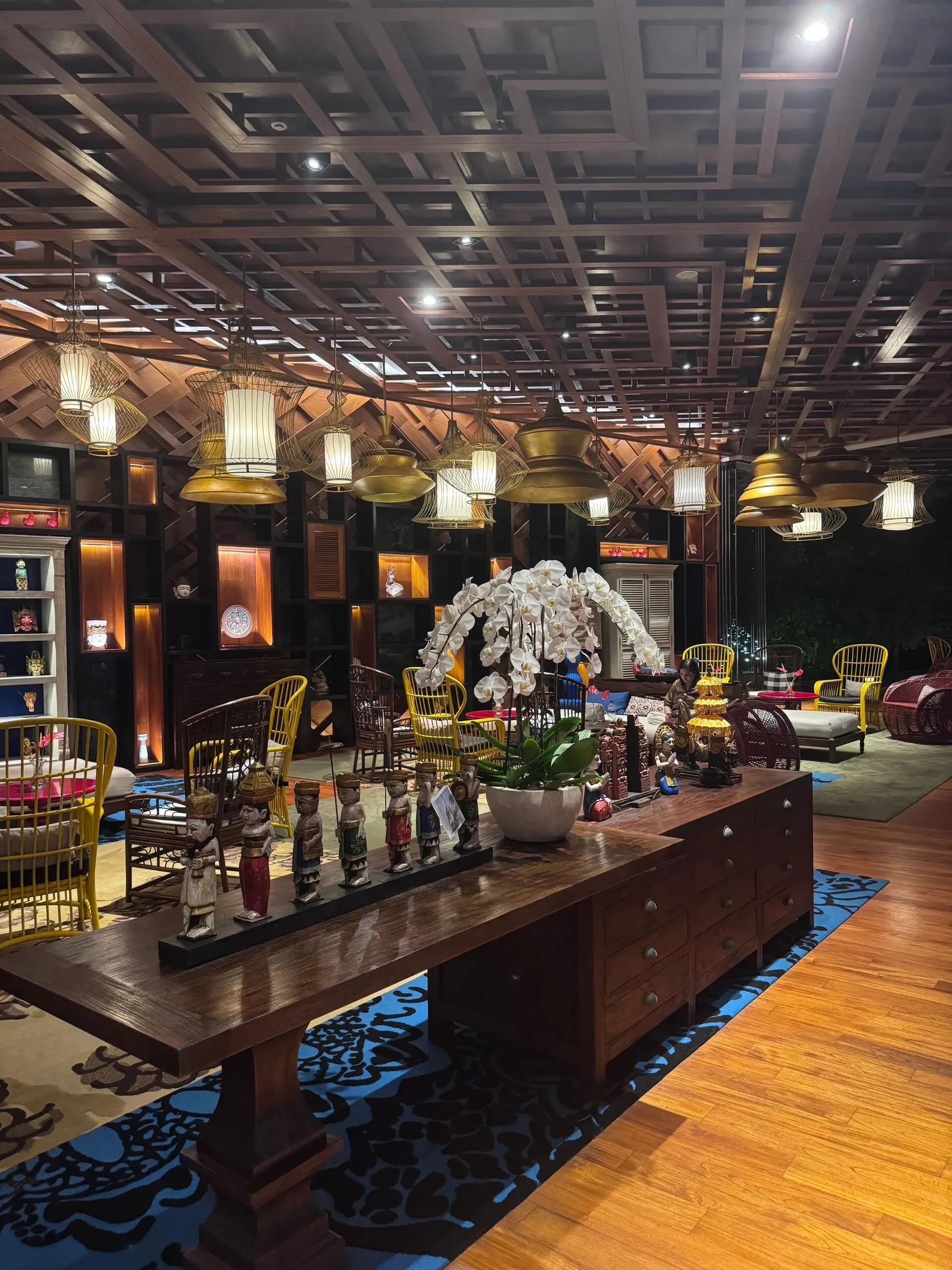 The lobby lounge at night, with lattice timber ceilings, brass lanterns, yellow rattan chairs and white orchids