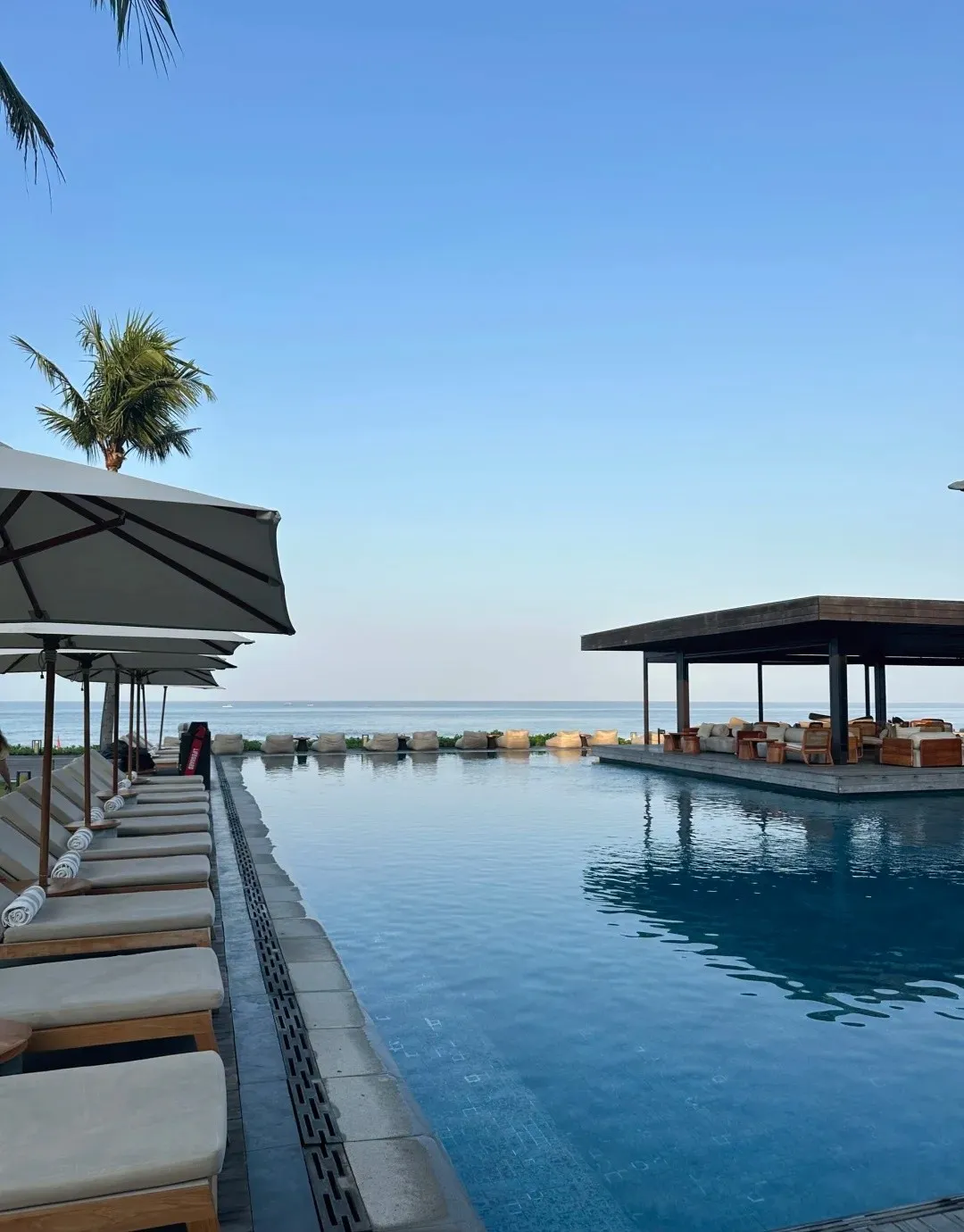 Sun loungers along the edge of a long pool, with a pavilion bar at the far end by the sea