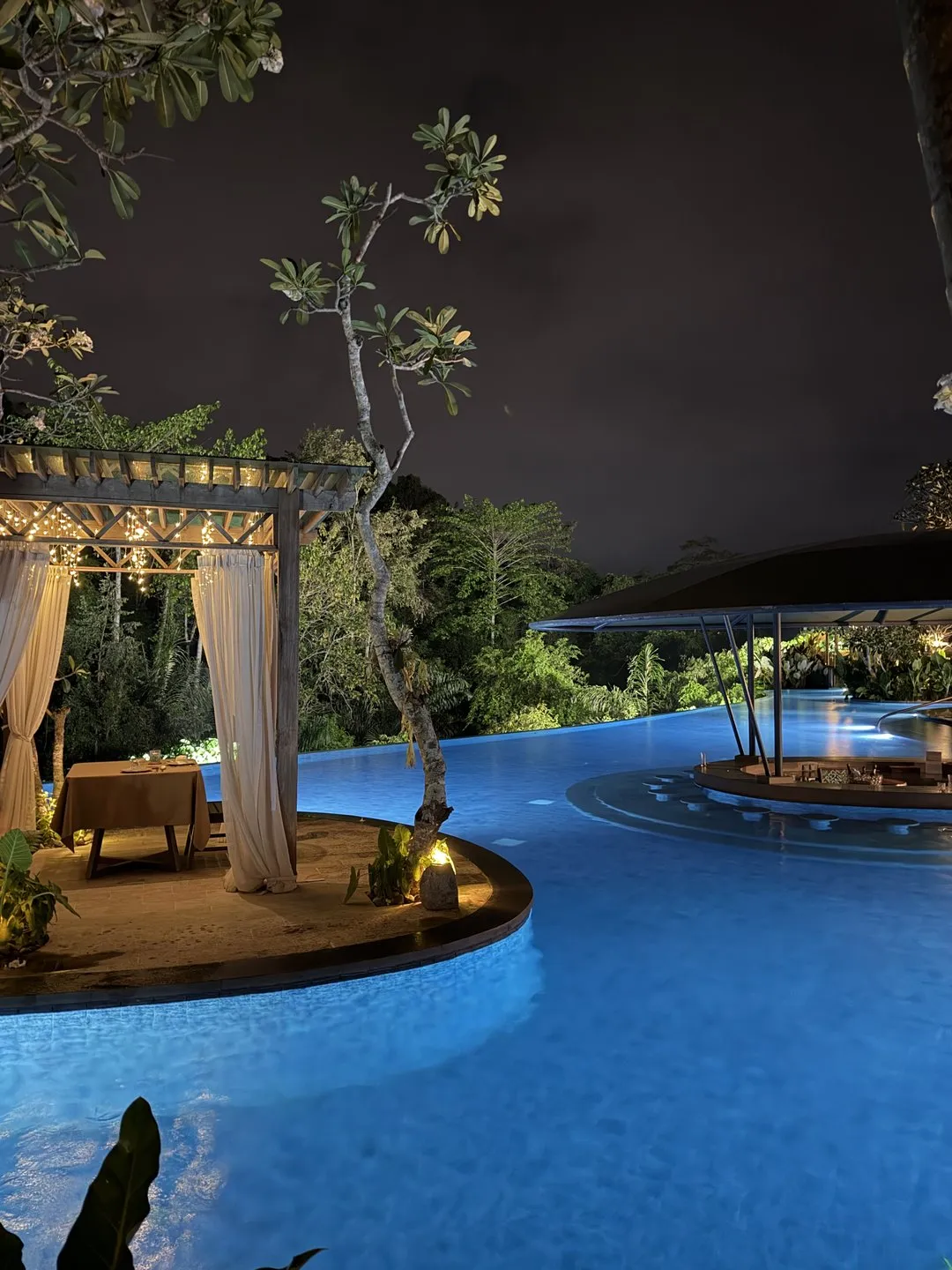 The pool lit blue at night, with a curtained pavilion on its own island