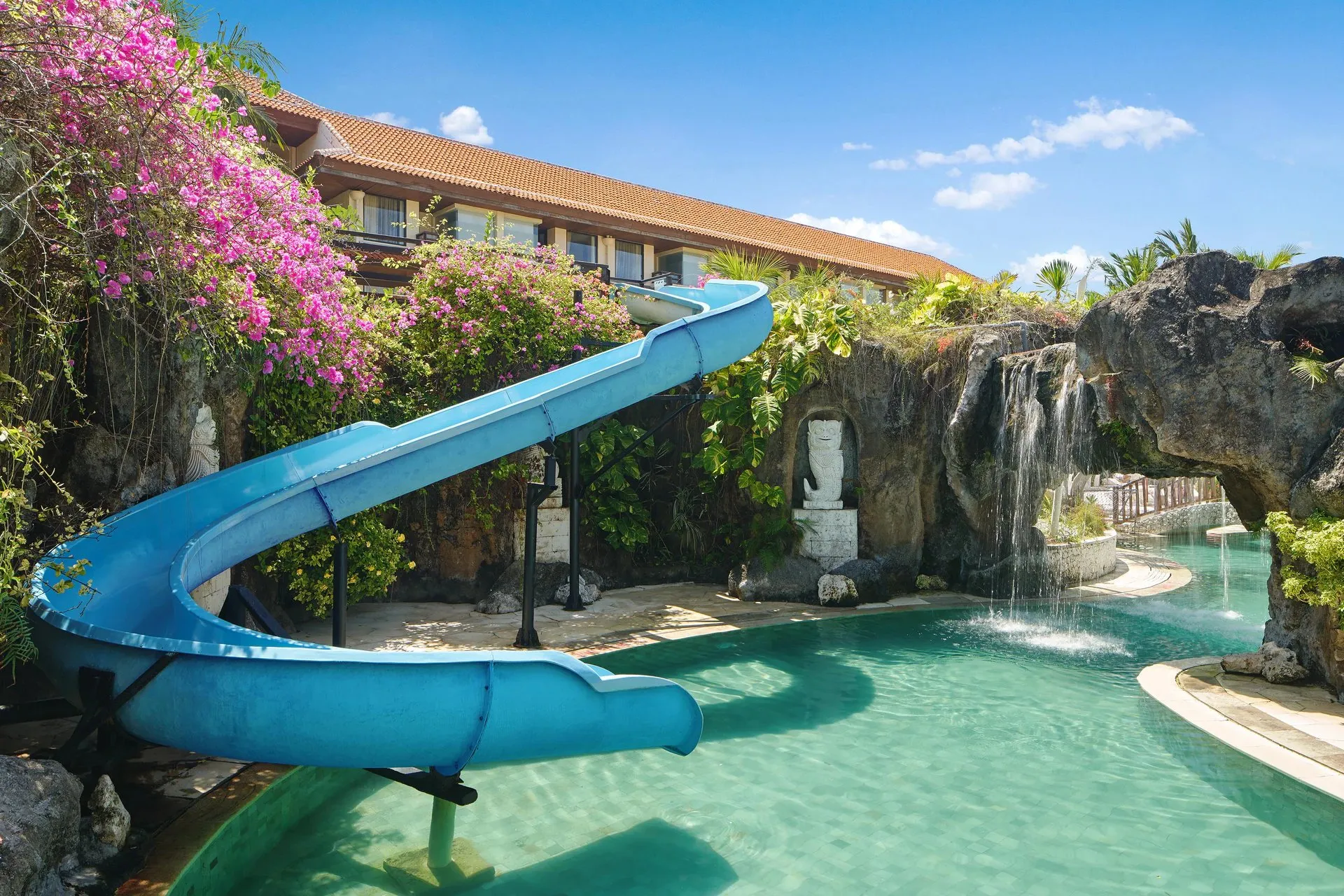 The blue water slide curving into the lagoon pool beside a rock waterfall, under bougainvillea