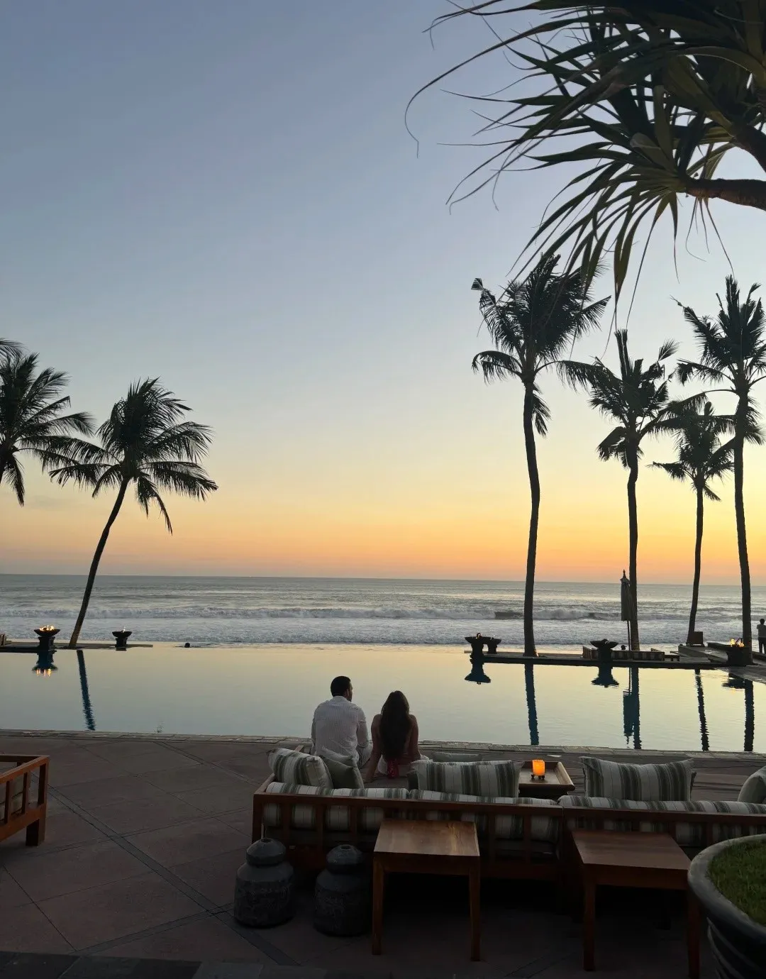 A couple on a sofa by the infinity pool at sunset, with palms silhouetted against an orange sky
