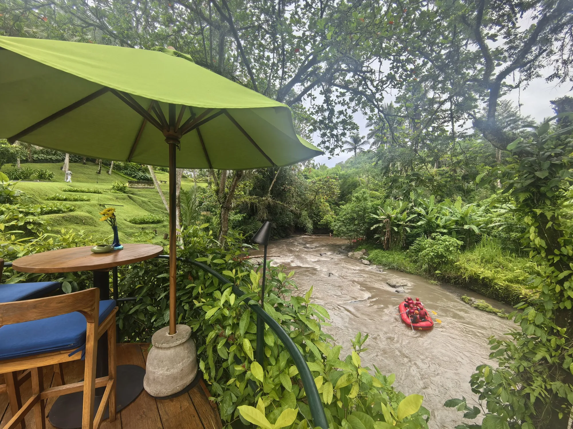 A table under a green umbrella above the Ayung River, with a red raft going past below