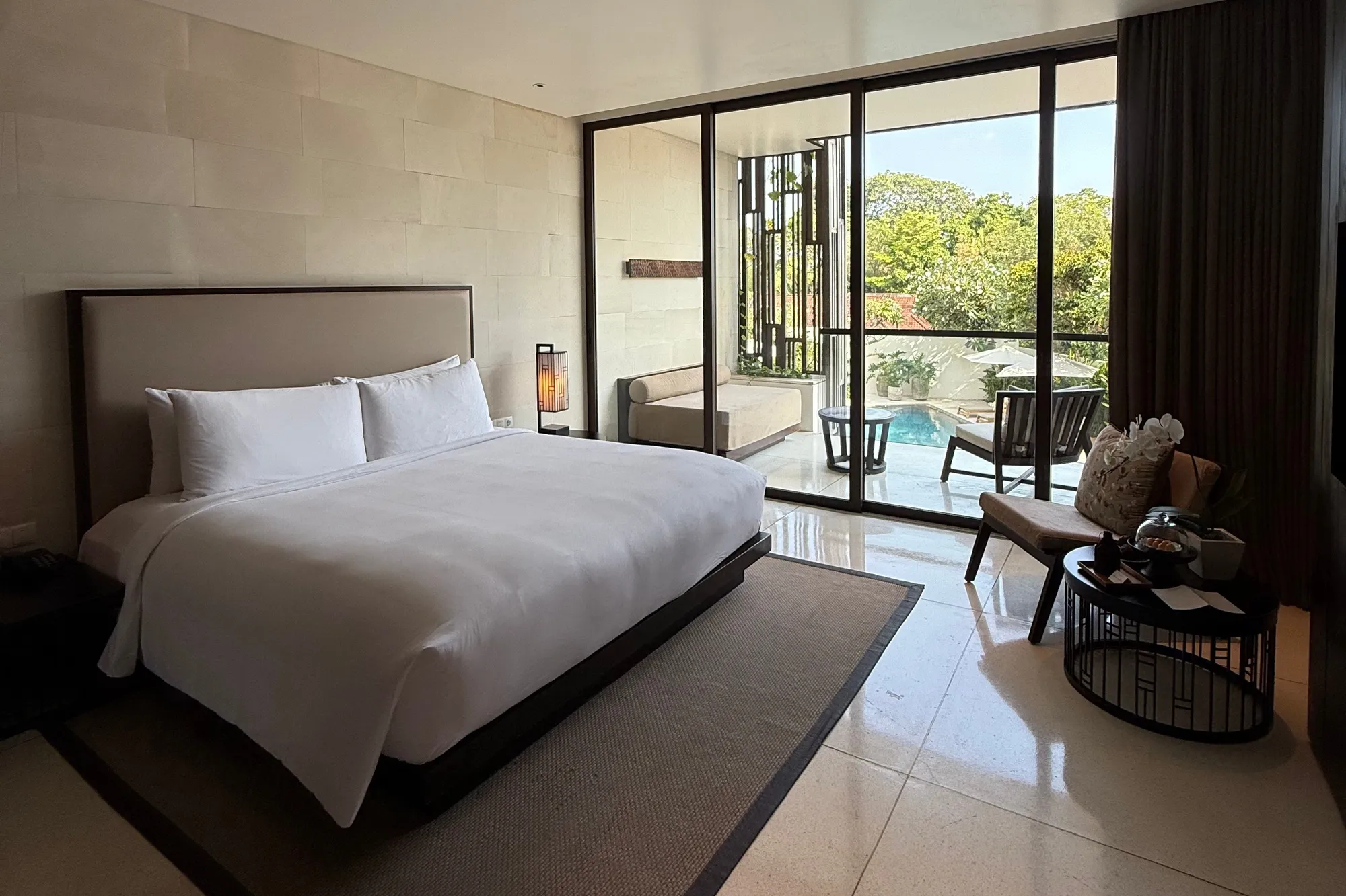 An Alila Seminyak bedroom with a king bed, stone walls and glass doors opening onto a terrace above a pool