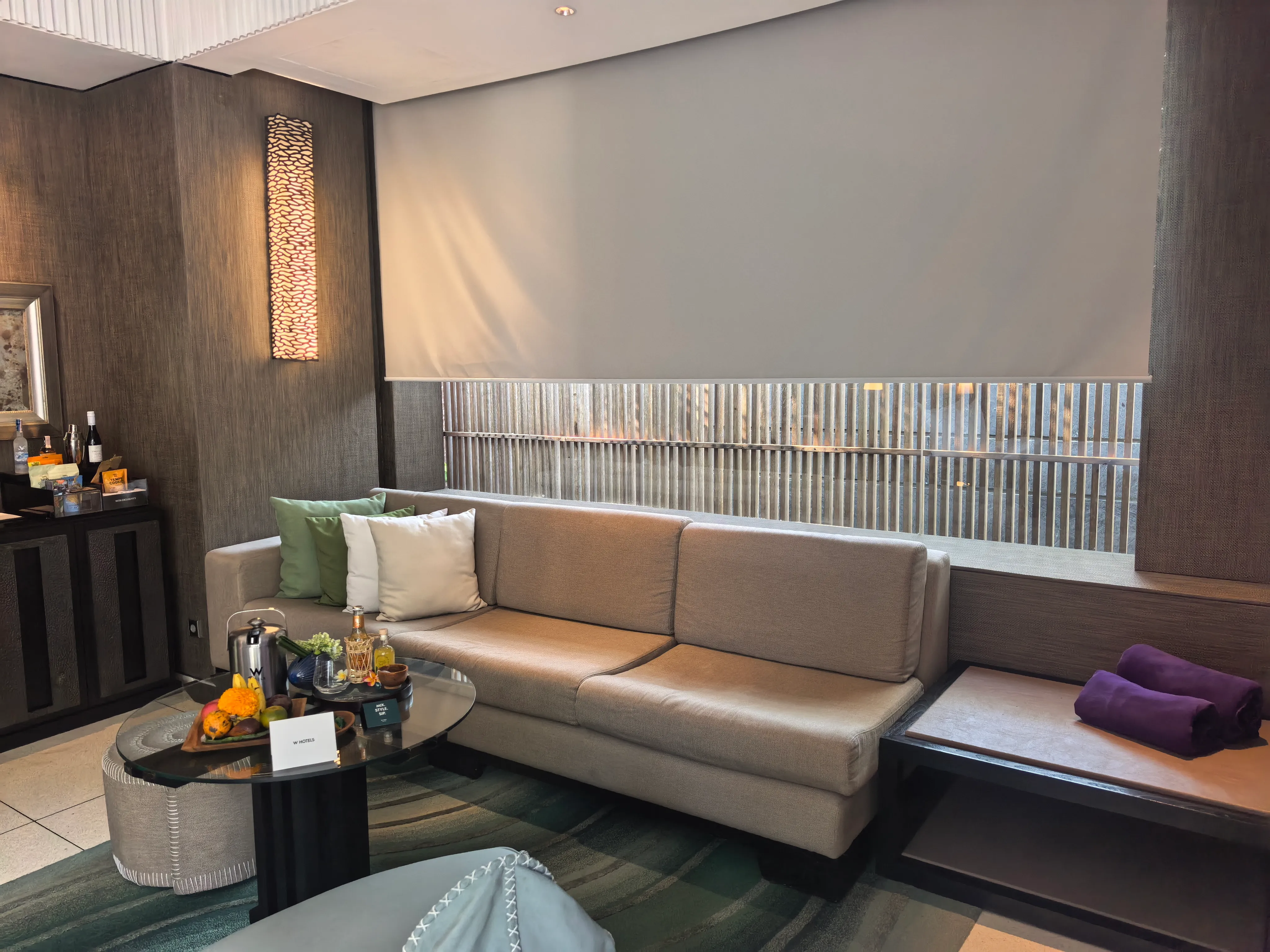 A villa living room with a long sofa under a slatted window, a fruit platter on the coffee table and a minibar