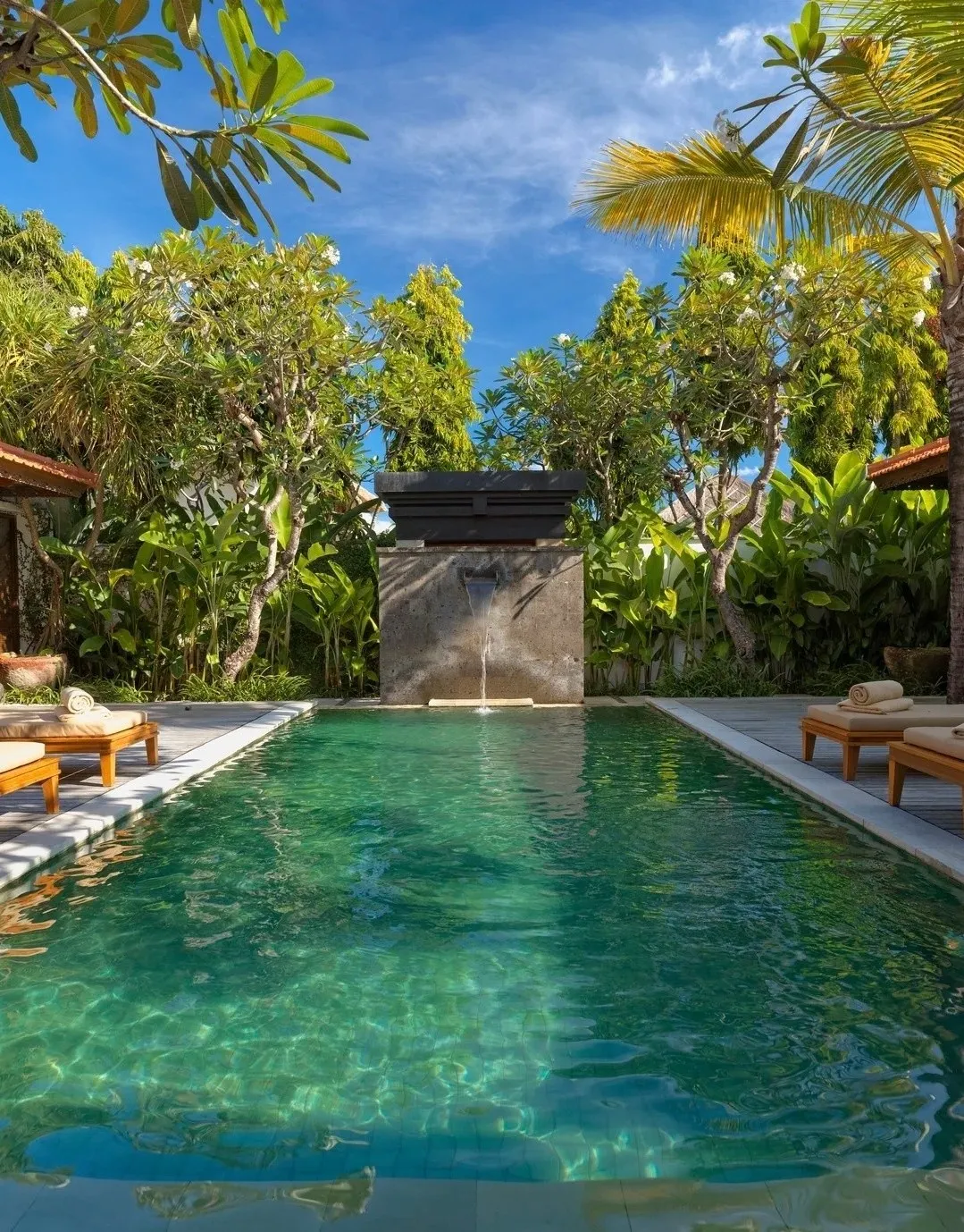 A long private villa pool between sun loungers, with a stone water spout and tropical garden behind
