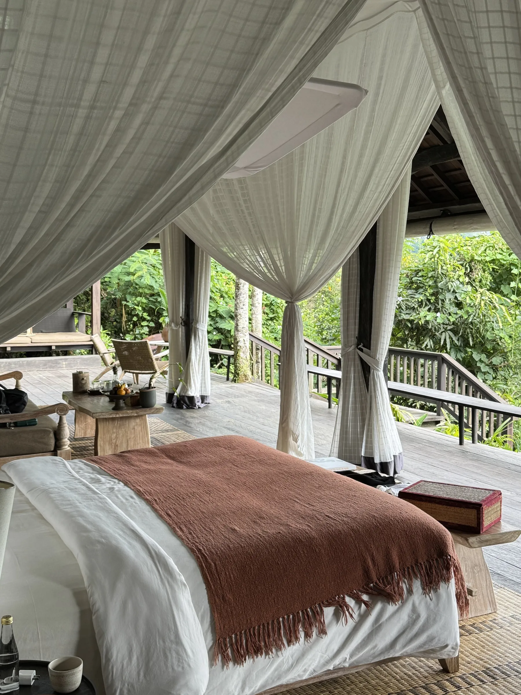 A bed under white canopy drapes in an open-air villa, with a timber deck and jungle all around and no walls in between