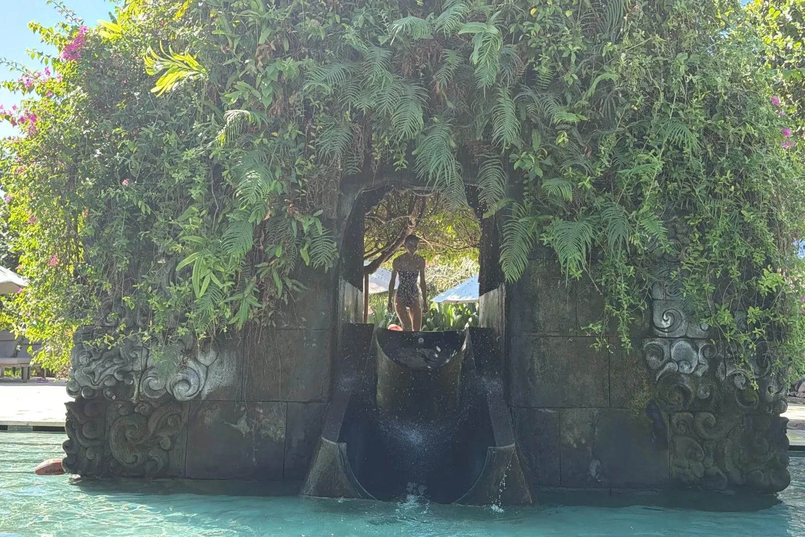 A water slide running out of a fern-covered, carved stone grotto into the pool